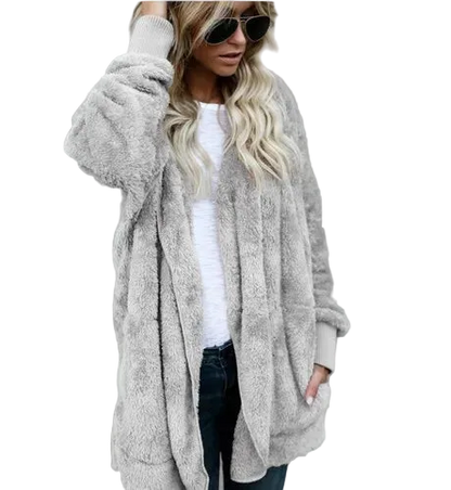 Woman wearing a long beige cardigan over a white top and dark jeans against a gray concrete wall.