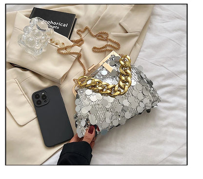 Silver sequin clutch with gold chain held by a hand on a white surface with a beige jacket and phone.